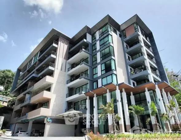 Modern multi-story building with sleek design and balconies, surrounded by lush greenery.