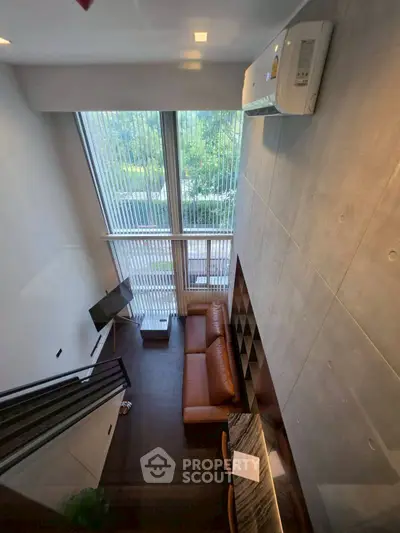 Modern loft-style living room with high ceilings and large windows offering a lush green view.