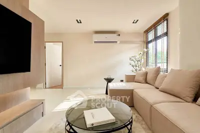 Modern living room with beige sofa, large TV, and natural light from window blinds.
