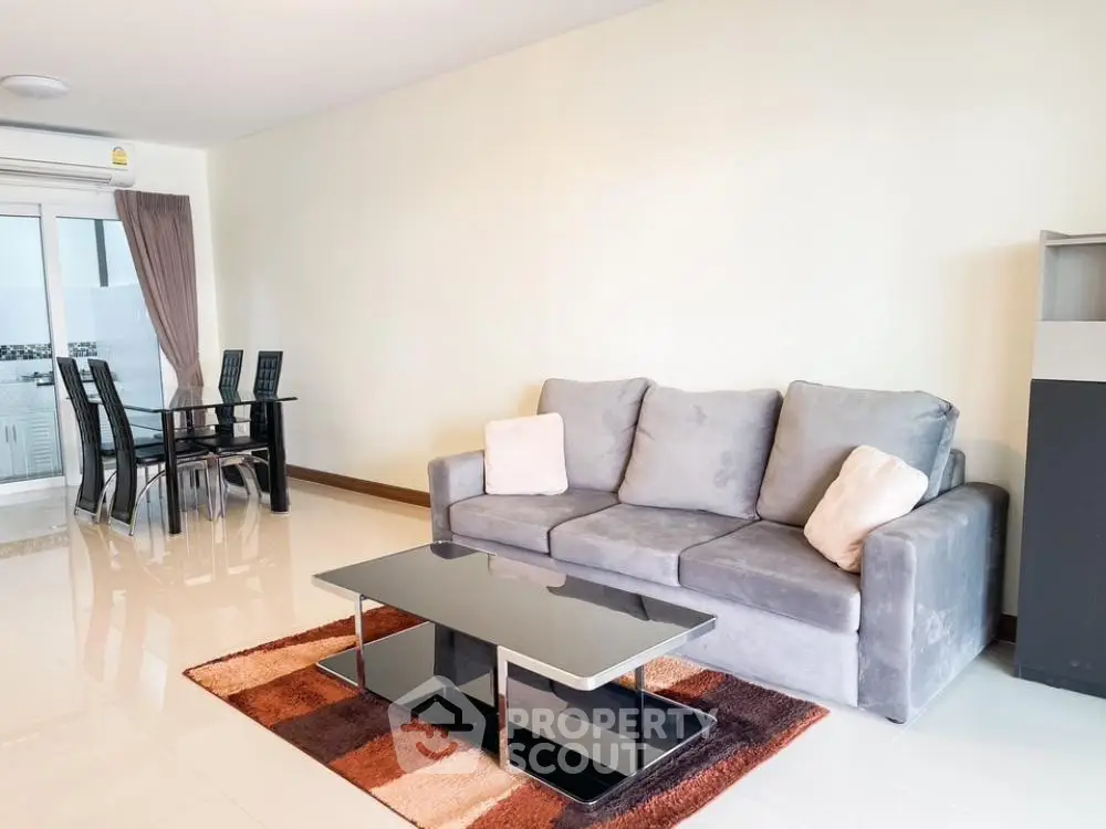 Spacious living room with glossy tiled floors, modern grey sofa, elegant glass coffee table, and a sleek dining set by the window, exuding contemporary charm.