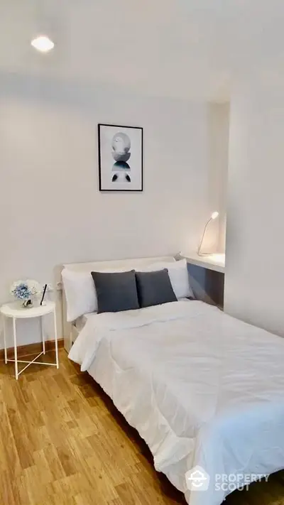 Cozy minimalist bedroom with polished hardwood floors and a comfortable double bed, accented by stylish gray pillows and a chic bedside table with a lamp.