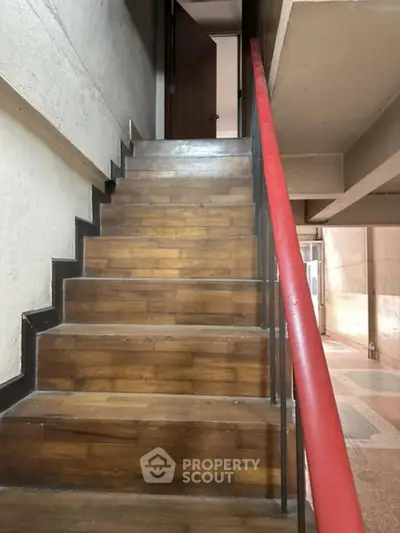 Elegant wooden staircase with a striking red handrail, leading to upper levels in a modern residential building, showcasing architectural detail and design.