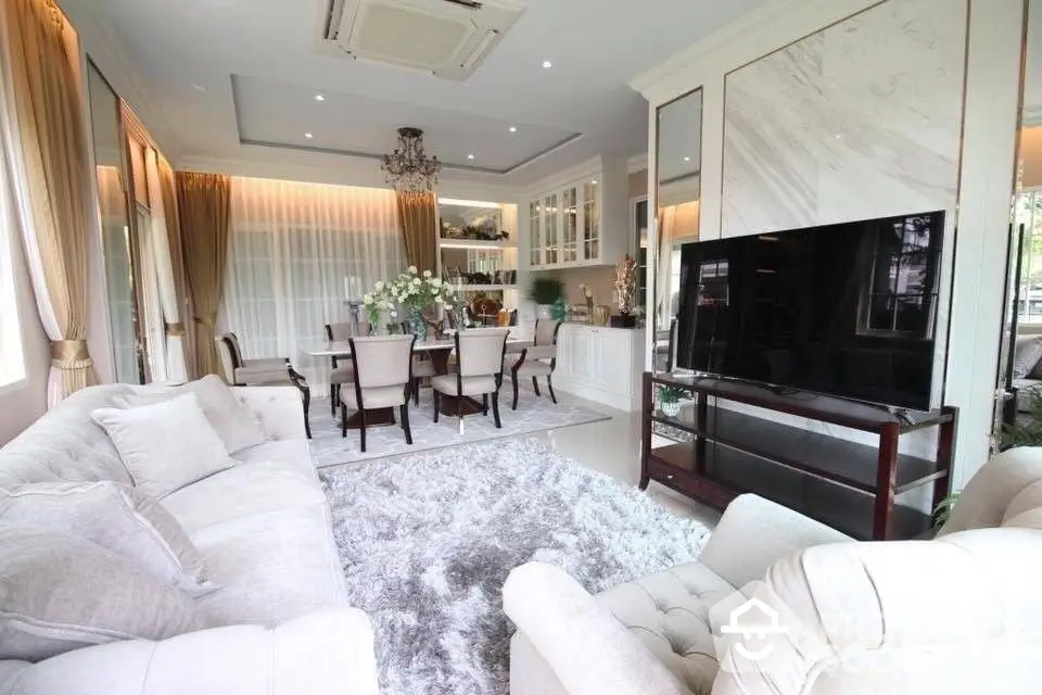 Elegant open-plan living space with plush white sofas, a large TV, and a dining area, bathed in natural light from large windows, perfect for entertaining.