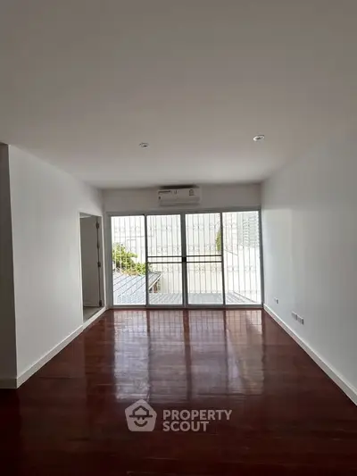 Spacious living room with polished wooden floors and large windows leading to a balcony.