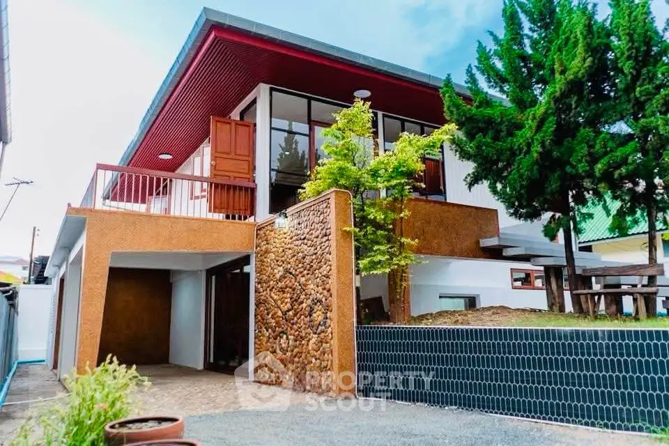 Modern two-story house with unique stone facade and lush greenery, featuring a spacious balcony and contemporary design.