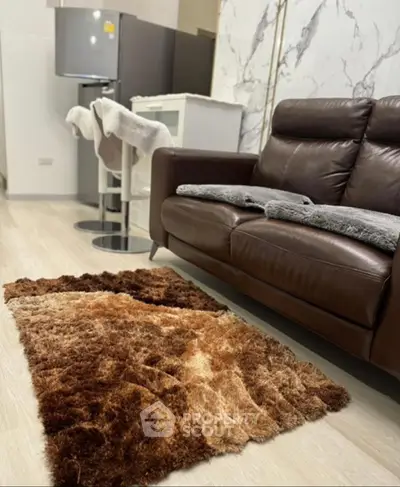 Modern living room with leather sofa and plush rug, featuring sleek kitchen appliances in the background.