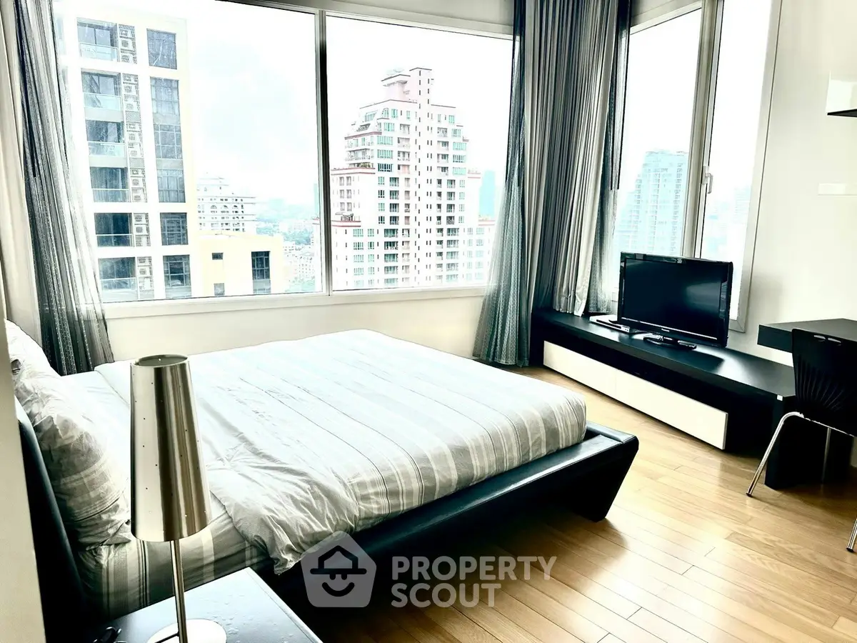 Modern bedroom with large windows and city view, featuring sleek furniture and wooden flooring.