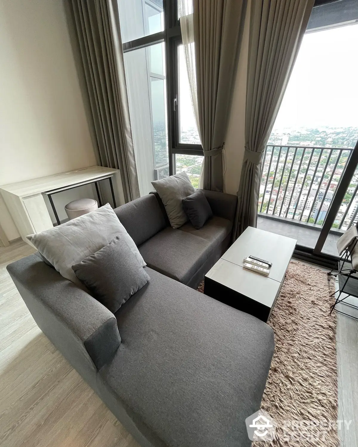 Cozy living room with modern grey sofa, plush area rug, and floor-to-ceiling windows offering a panoramic city view.