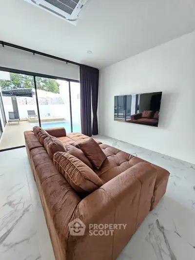 Modern living room with plush brown sofa, wall-mounted TV, and large glass doors leading to a patio.