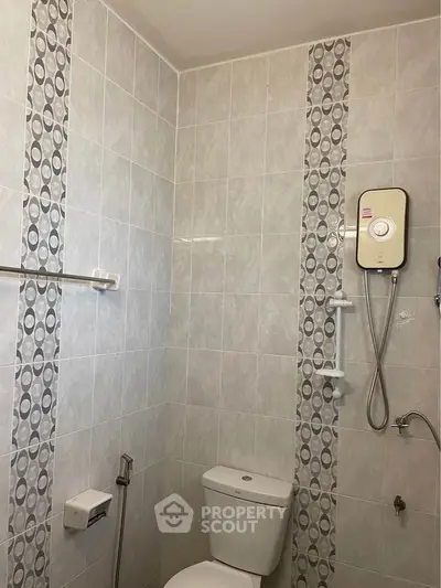 Compact bathroom with modern electric shower unit and neutral tile design, featuring a clean white toilet and convenient safety grab bars.