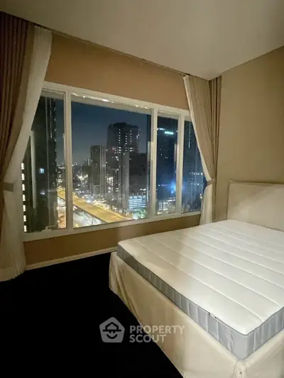 Spacious bedroom with large window offering stunning city skyline view at night.