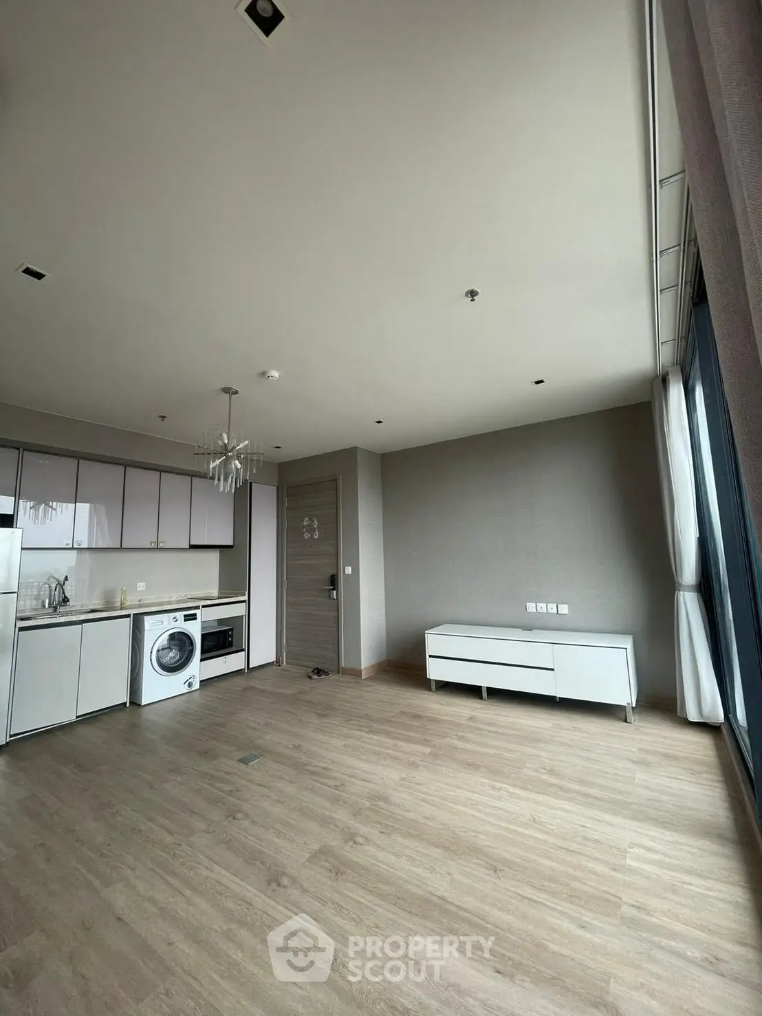 Modern open-plan living area with kitchen and washing machine, featuring large windows and wooden flooring.