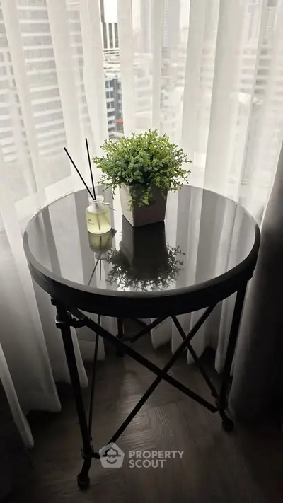 Chic round table with plant and candle by window, offering urban view.
