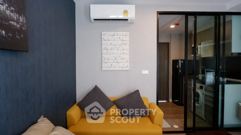 Chic modern living space with a vibrant yellow sofa, sleek air conditioning unit, and a view into a well-equipped kitchen with modern appliances.