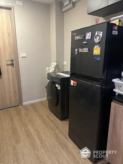 Modern kitchen with sleek black fridge and washing machine, wooden flooring, and minimalist design.