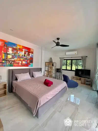 Spacious bedroom with polished tile flooring, modern ceiling fan, vibrant wall art, and ample natural light streaming through large windows, creating an inviting and comfortable living space.