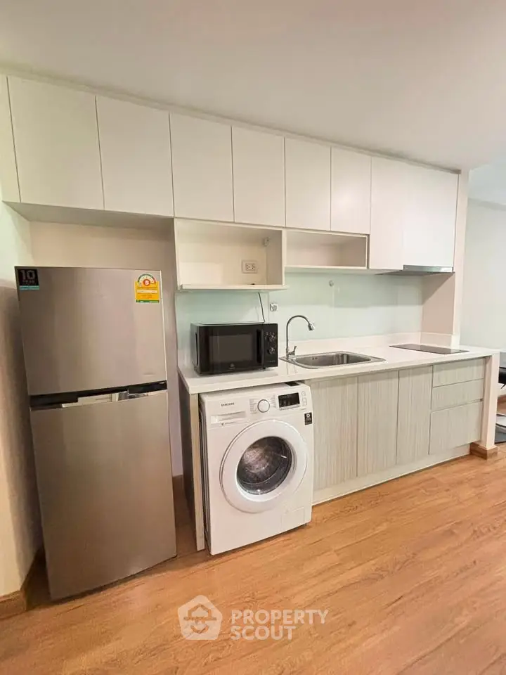 Modern kitchen with sleek cabinets, stainless steel fridge, washing machine, and microwave on wooden flooring.