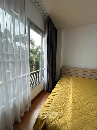 Cozy bedroom with large window and yellow bedding, offering a serene view of lush greenery.