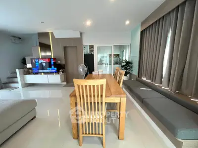 Modern dining area with wooden table and chairs, adjacent to a cozy living space with a TV and large windows.