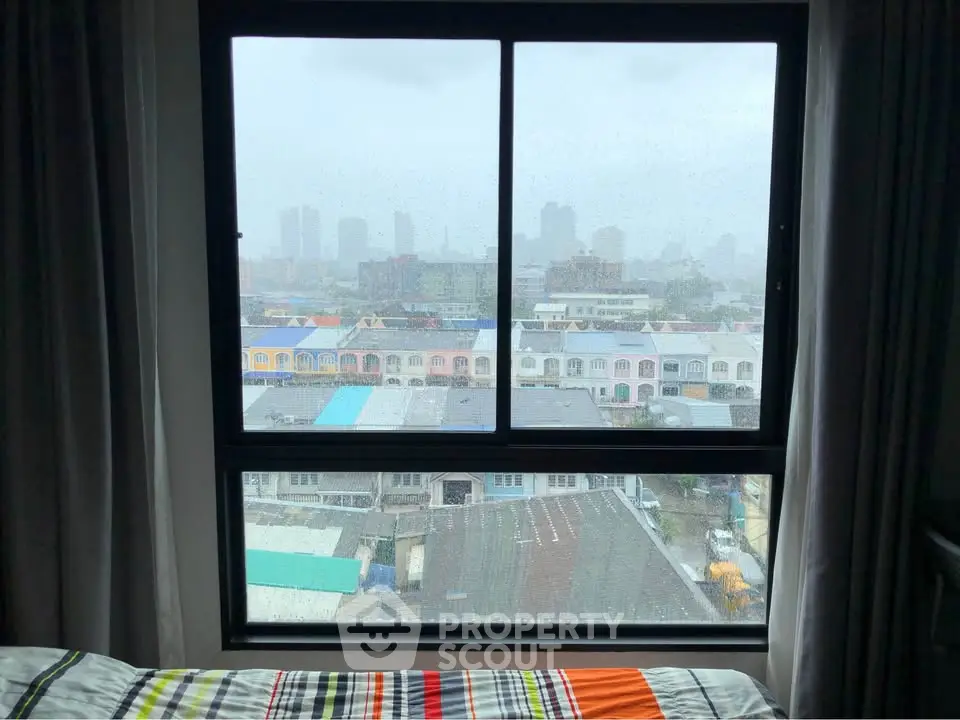Cozy bedroom with large window offering cityscape view on a rainy day.