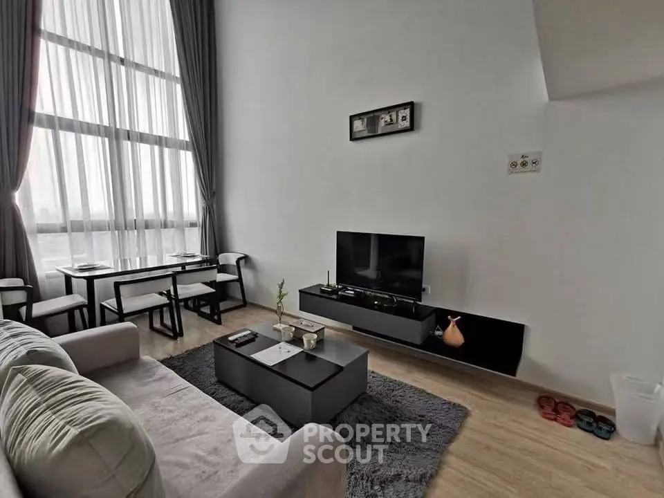 Modern living room with high ceilings and large windows, featuring stylish furniture and a cozy atmosphere.