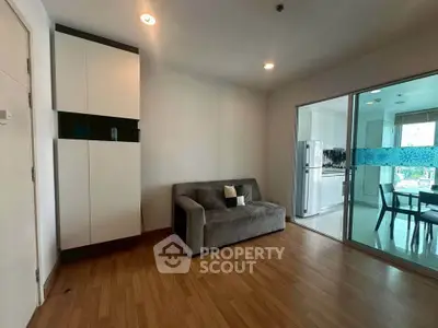 Modern living room with wooden flooring and glass sliding doors leading to a bright kitchen area.