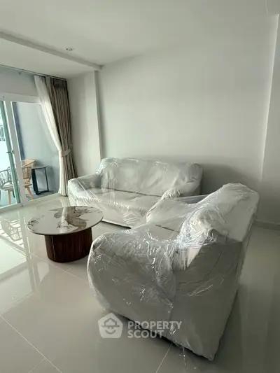 Modern living room with new furniture wrapped in plastic, featuring a sleek coffee table and balcony access.