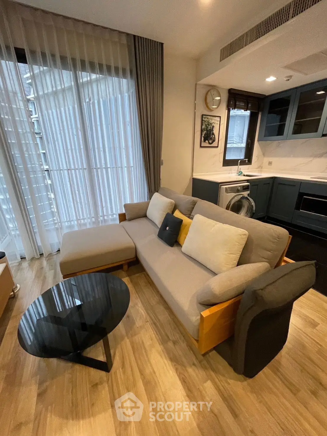 Modern living room with stylish sofa and open kitchen layout, featuring floor-to-ceiling windows and wooden flooring.