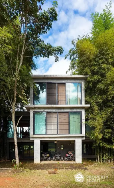 Modern three-story house with large windows surrounded by lush greenery and trees.