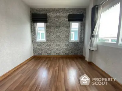 Spacious room with polished wooden floors and a stylish brick accent wall flanked by two large windows inviting ample natural light.