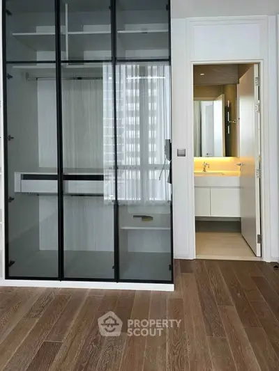 Modern apartment interior with glass closet and bathroom view, featuring sleek design and wooden flooring.
