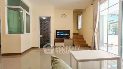 Modern living room with staircase and TV, featuring large windows and neutral decor.