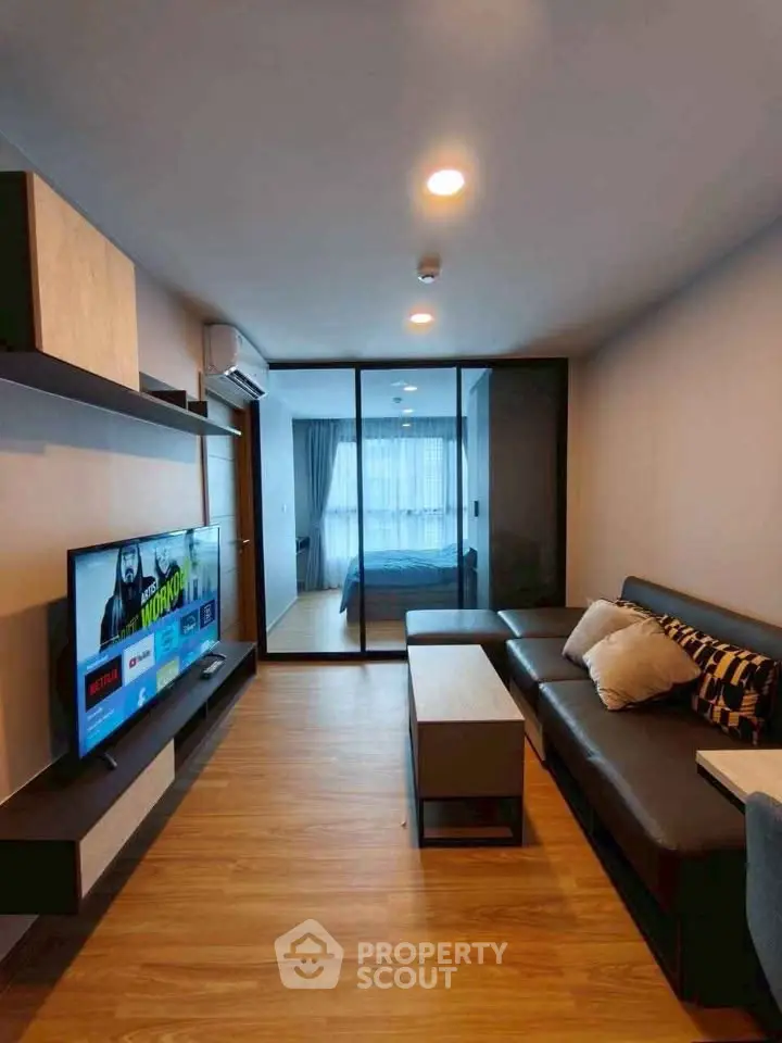 Modern living room with sleek furniture and TV, leading to a cozy bedroom through glass doors.