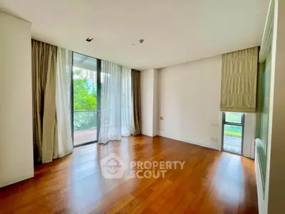 Spacious empty room with hardwood floors and large windows opening to a balcony with lush greenery view.