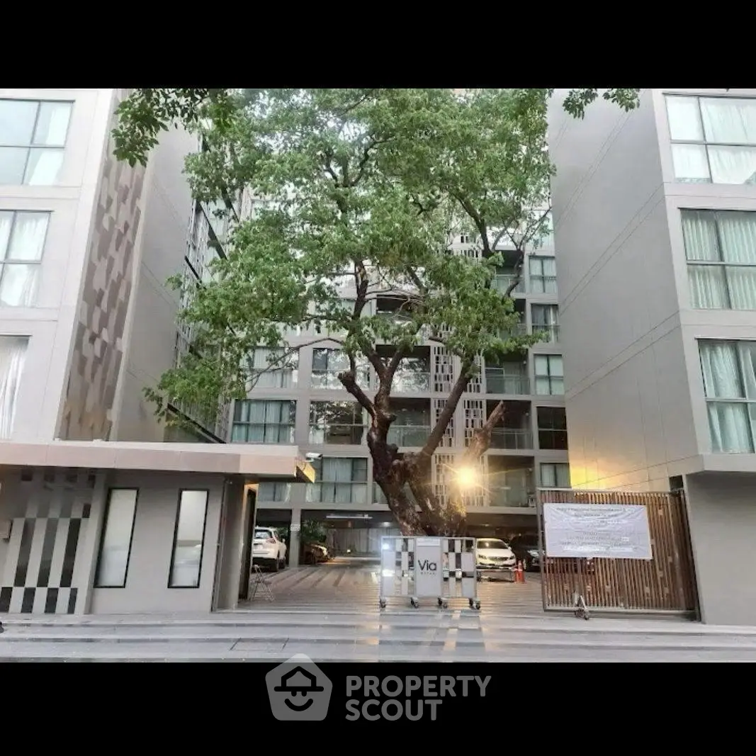 Modern apartment building entrance with lush tree and parking area.