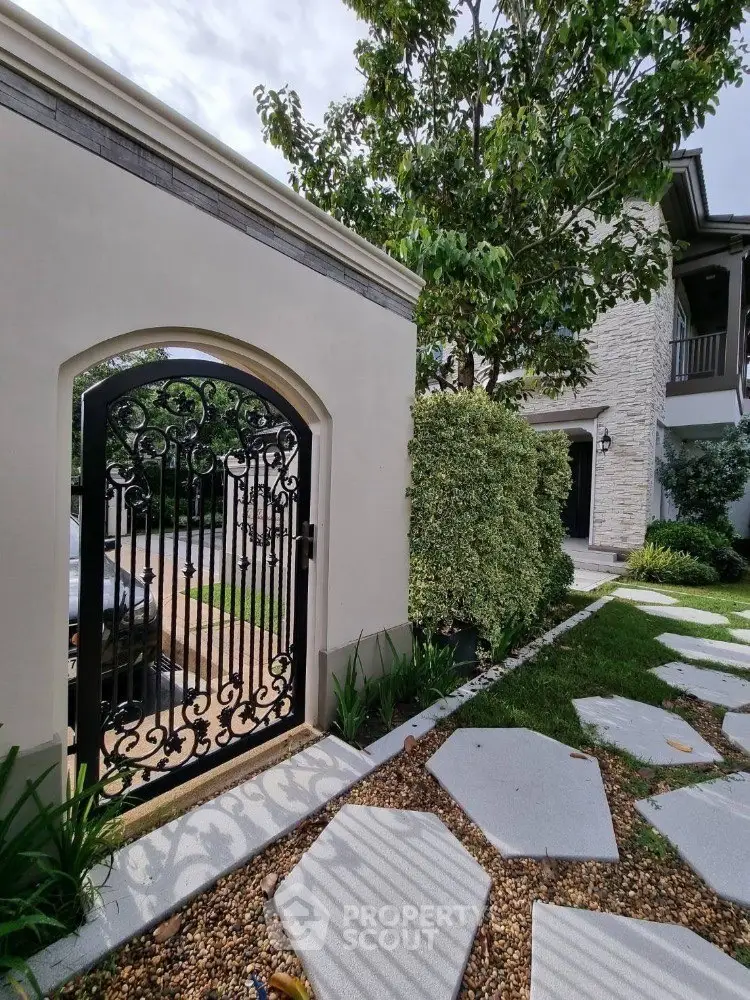 Charming garden entrance with elegant wrought iron gate and lush greenery, leading to a beautiful stone-clad house.