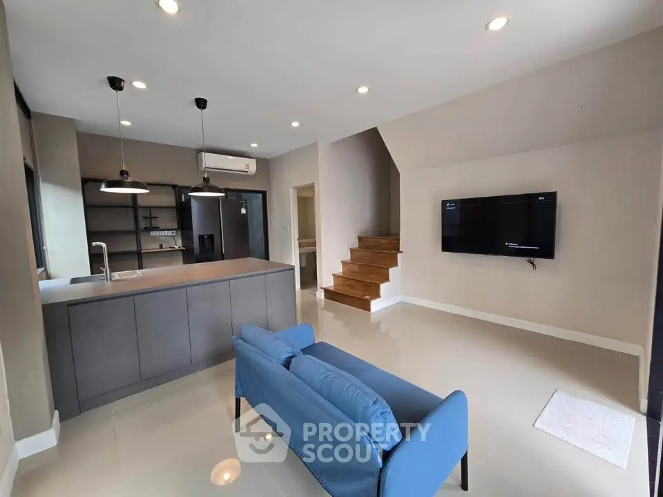 Modern open-plan living room with sleek kitchen and stylish staircase, featuring a cozy blue sofa and wall-mounted TV.