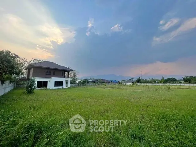 Expansive green field with a distant view of a house and mountains under a dramatic sky.
