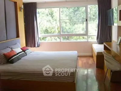 Bright bedroom with large window and wooden flooring, offering a serene view of lush greenery.