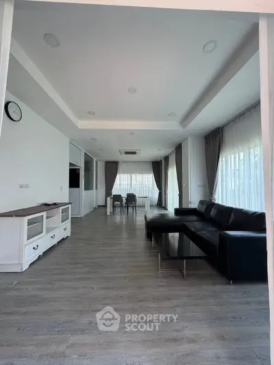 Spacious modern living room with sleek black sofa and elegant dining area, featuring large windows and stylish flooring.