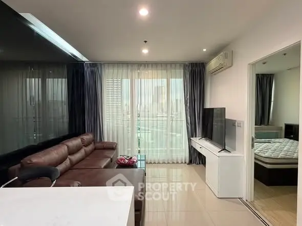 Modern living room with leather sofa and large TV, adjacent to a bedroom with natural light.