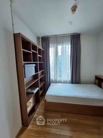 Cozy bedroom with wooden flooring and built-in shelving, featuring a large window with curtains.