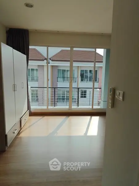 Bright room with large windows and wooden flooring, overlooking neighboring buildings.
