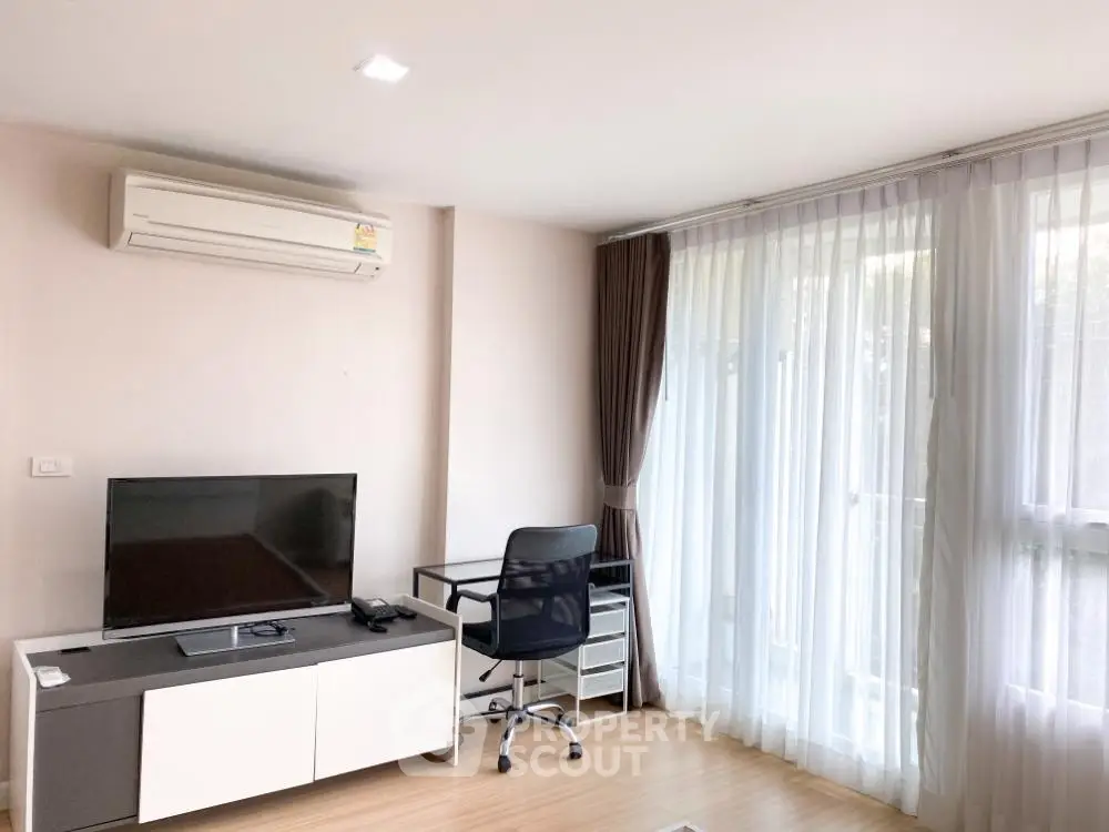 Spacious and well-lit living room with modern furnishings, large windows draped with sheer curtains, and a sleek entertainment unit.