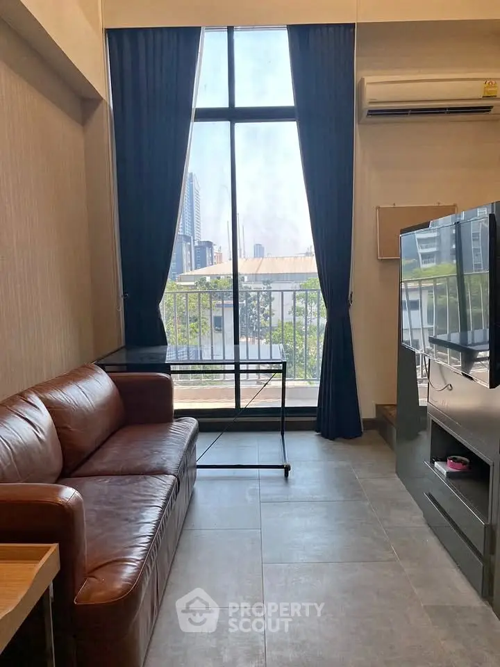 Modern living room with large window and city view, featuring a leather sofa and sleek TV setup.