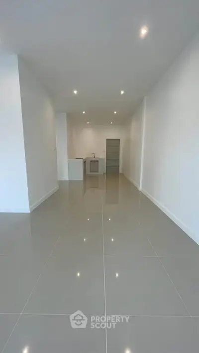 Spacious modern living area with sleek tile flooring and recessed lighting, leading to a compact kitchen.