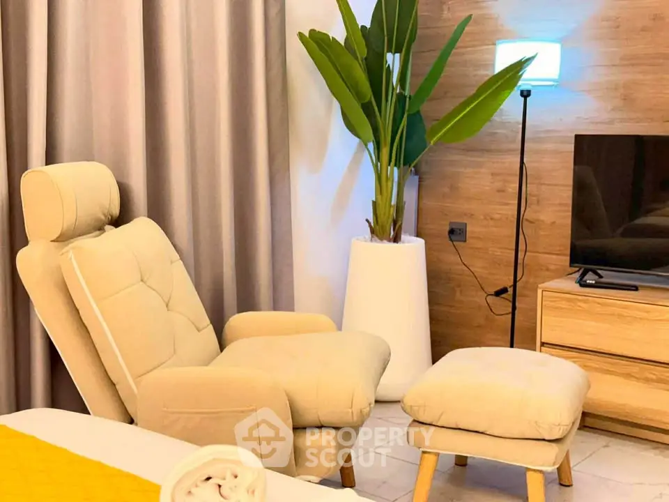 Cozy living room with modern beige armchair, footrest, and TV setup, accented by a tall plant and stylish lighting.