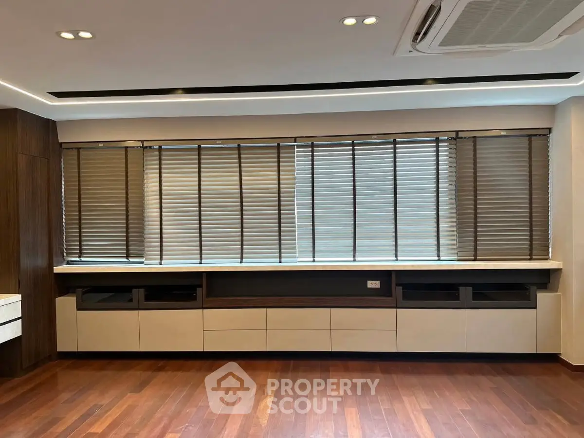 Modern living room with wooden flooring and large windows with blinds, featuring built-in storage units.