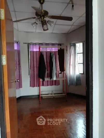 Cozy bedroom with wooden flooring and ceiling fan, featuring a clothes rack and purple curtains.