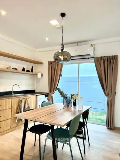Modern kitchen with dining area, featuring sleek wooden cabinets, stylish lighting, and a large window with curtains.